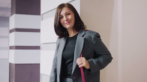 smiling confident businesswoman with red handbag standing against wall in office corridor, looking back over shoulder, modern office environment, stylish gray suit, professional appearance