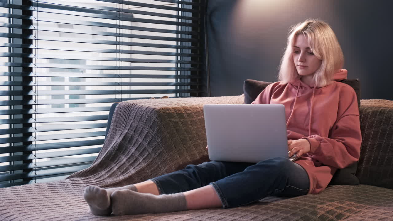 Young content creator blonde girl is on her laptop sitting on the sofa near the window. Working from home