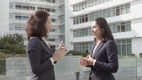 two smiling business ladies standing outside office building, talking and gesturing with hands