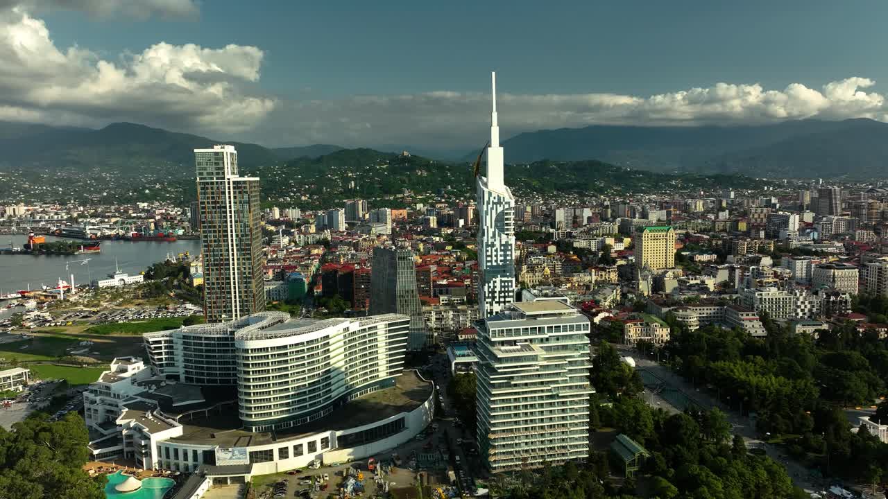 A panoramic aerial view of a coastal city featuring a tall architectural landmark and surrounding urban structures under partly cloudy skies