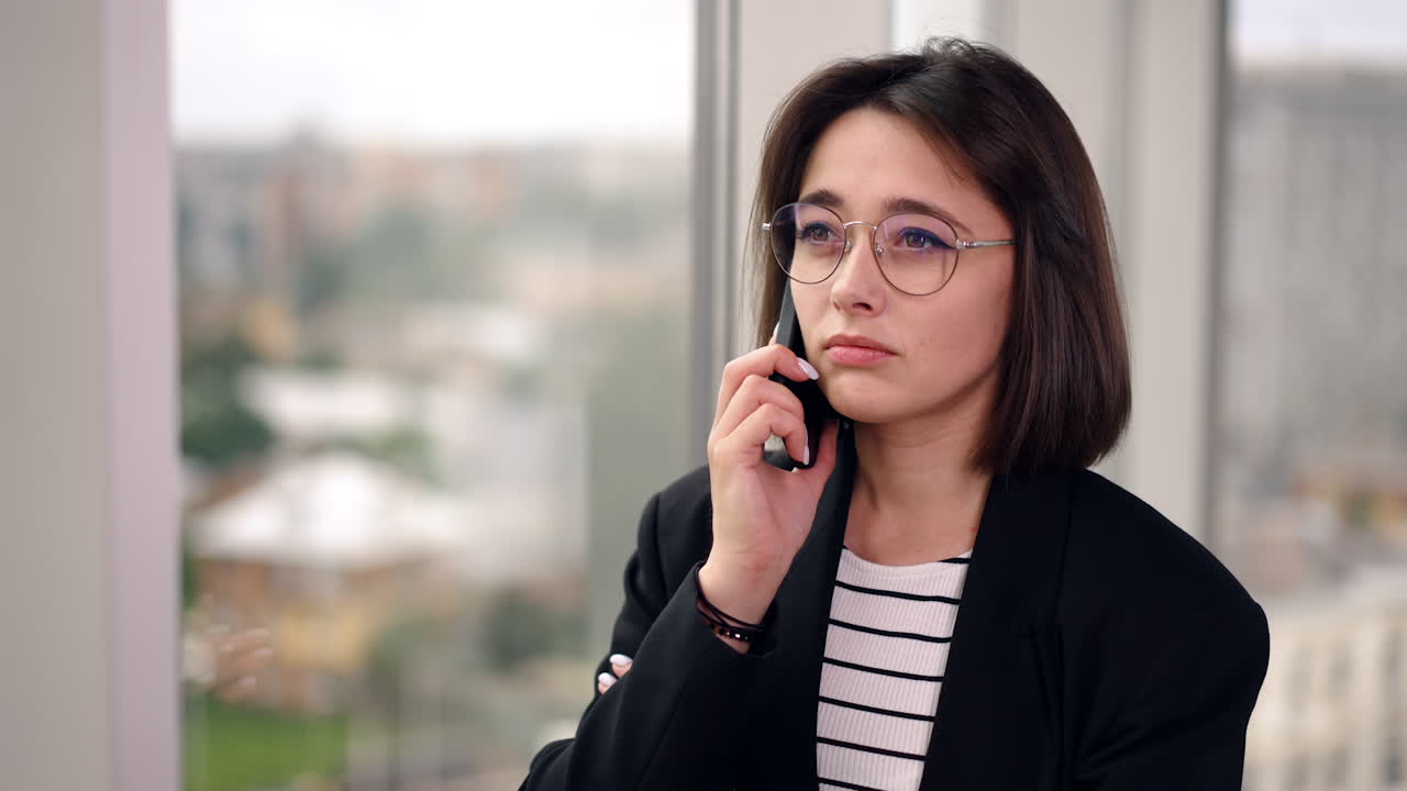 Caucasian woman wearing glasses is frowning as she speaks on the phone. In a moment lady cheers up smiling and looking at the window.