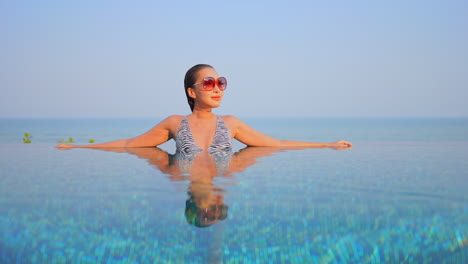 Mujer-Joven-Asiática-Apoyada-En-El-Borde-Dentro-De-La-Piscina-Infinita-Vista-Frontal-Cielo-Despejado-Durante-El-Día,-Espacio-De-Copia-De-Plantilla