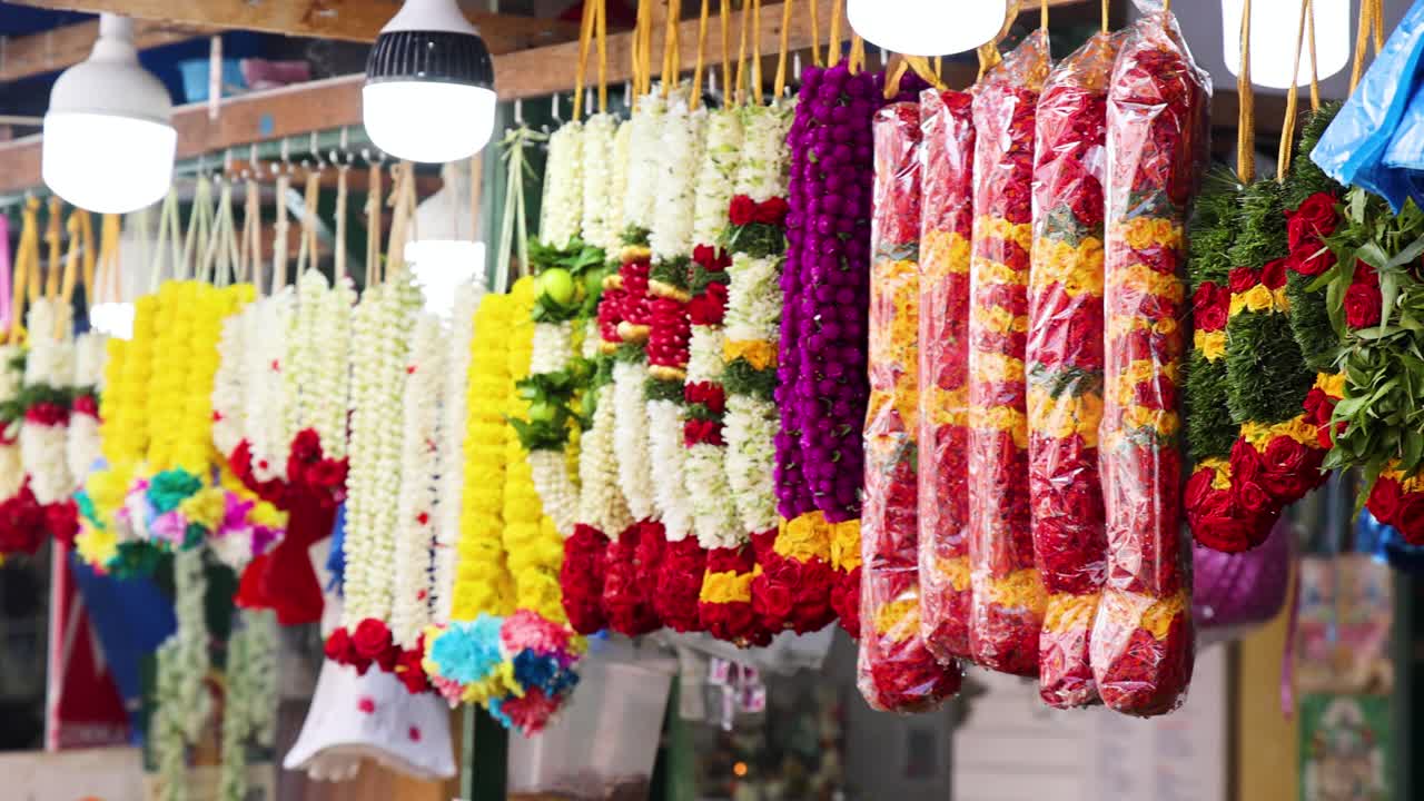 Rows of vibrant flower garlands hang under bright lights in a bustling Singapore market, with a slow camera pan revealing intricate floral arrangements
