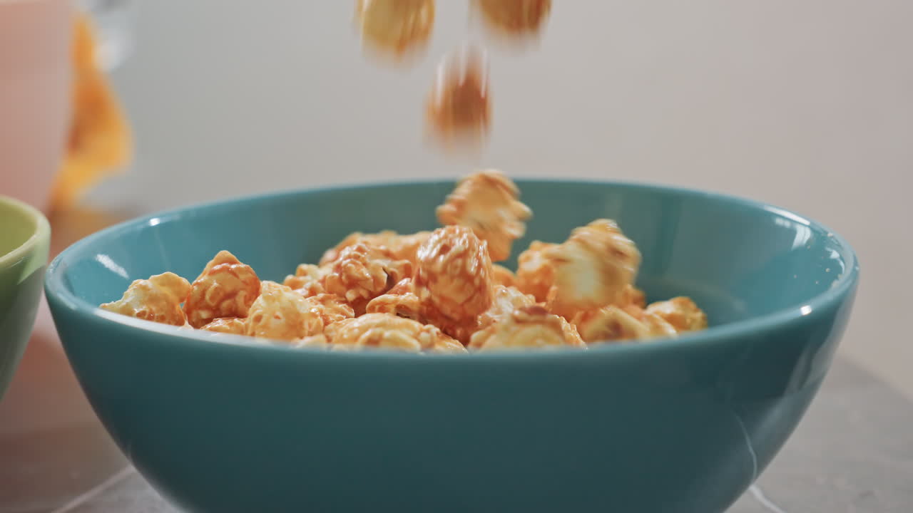 Close up view of honey flavored popcorn pouring from packet into blue bowl on table with green bowl filled with colorful candy visible in background, snack preparation with vivid colors and texture