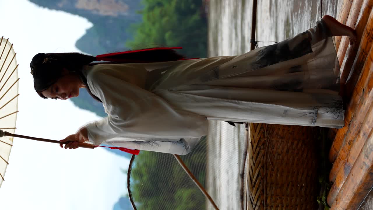 Woman in Hanfu attire holds an umbrella while standing on a bamboo raft in Xingping, China