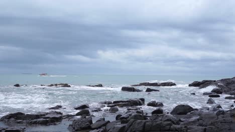 slowmo - ocean view with black rocks, waves, boat and seagull in bluff, new zealand on a cloudy day