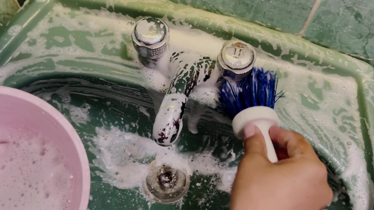 Primo piano di una mano che pulisce il lavello della cucina con spugna e acqua saponata, mostrando rubinetto, scarico, schiuma di sapone e superficie bagnata durante l'attività di pulizia domestica