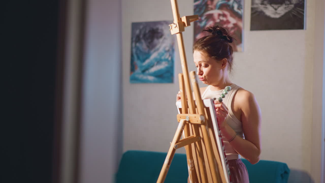 Young White Woman Painting At Easel, Soft Evening Light, Focused Expression, Holding Palette And Brush, Floral Canvas On Wooden Easel, Cozy Studio Interior, Warm Tones, Meditative Atmosphere