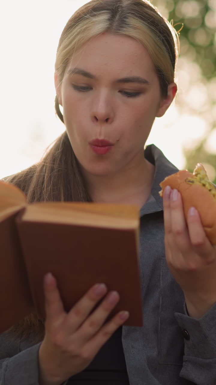 dama de camiseta gris comiendo bocadillo mientras lee un libro al aire libre, sentada en un banco del parque rodeada de árboles y vegetación con fondo borroso, la luz solar natural ilumina suavemente la escena
