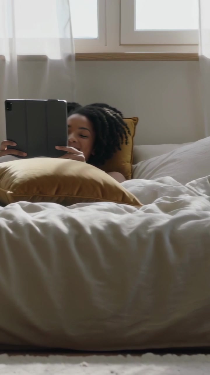 Vertikal video: Kvinde tapper på tablet, iført mørk top, læser i sengen ved vinduet og vender sig væk.
