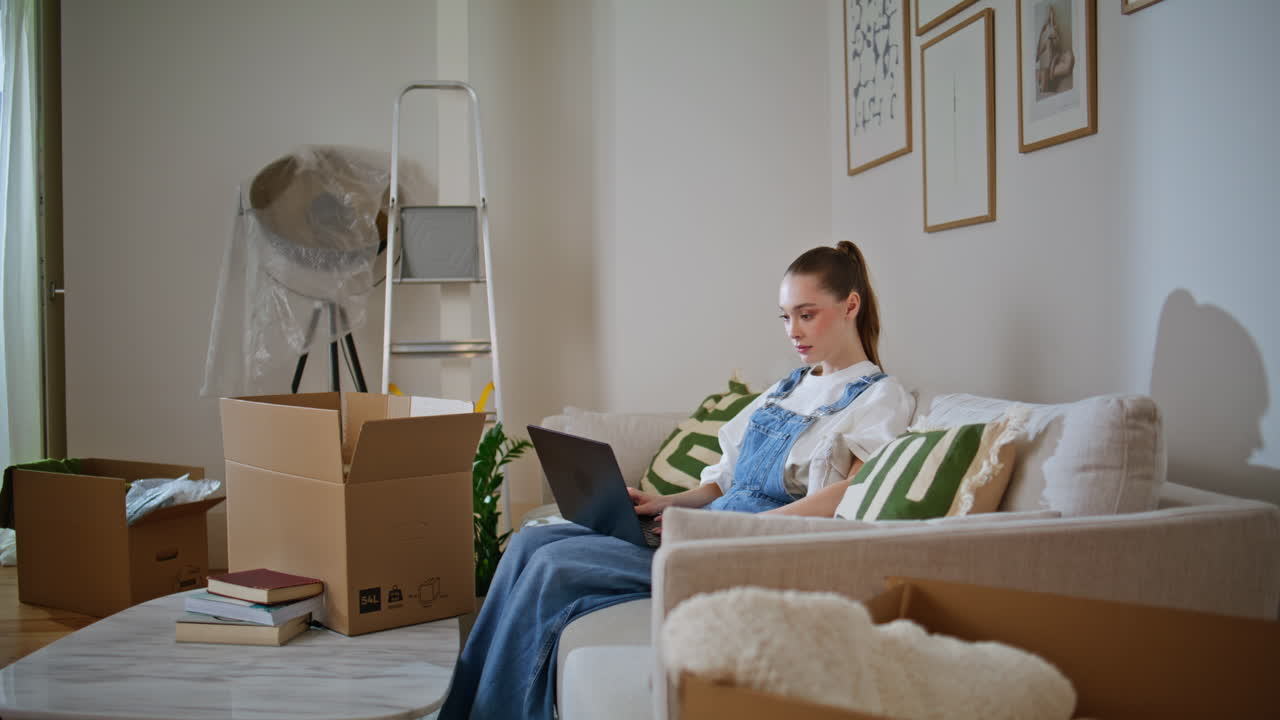 New apartment owner browsing laptop sitting couch near moving boxes. Woman home