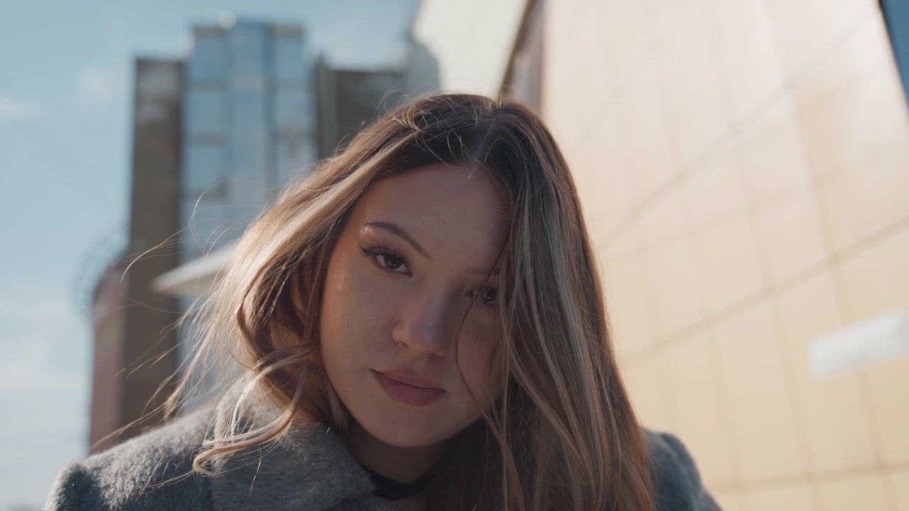 Bella giovane donna all'aperto con lo skyline della città, luce solare soffusa sui capelli e sul cappotto di lana, che guarda in basso con un sorriso gentile. Atmosfera urbana con uno sfondo di edificio in vetro riflettente, una leggera brezza che solleva ciocche