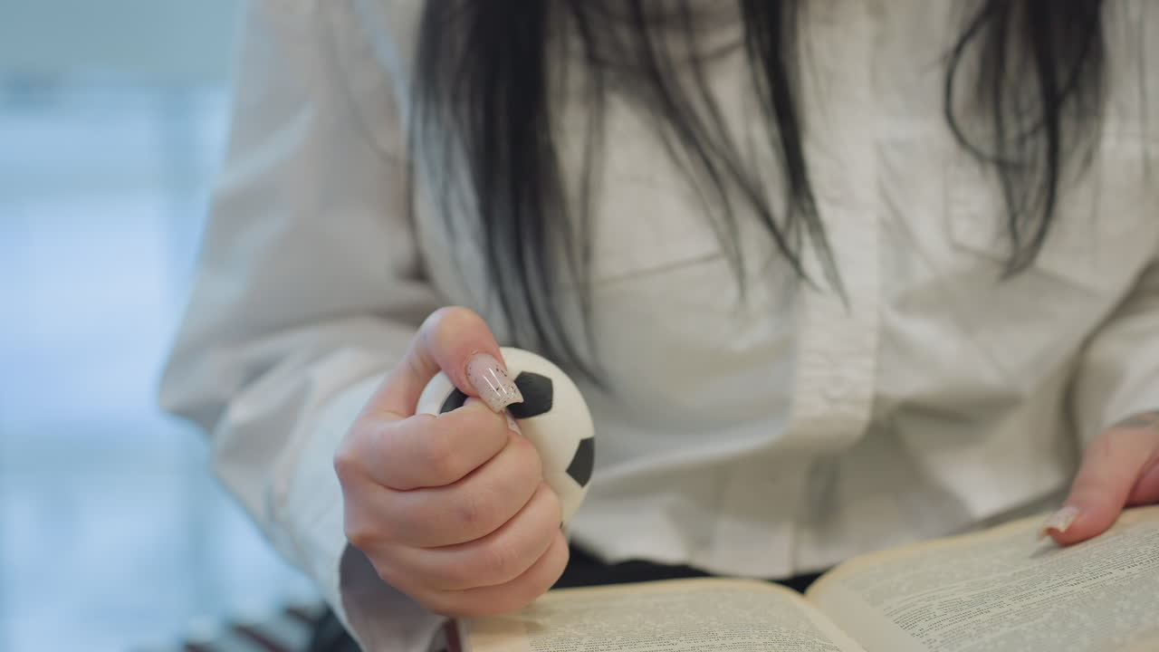 Close up of lady with long artificial polished nails squeezing black and white stress ball while reading, seated indoors with blurry moving cars seen through glass background
