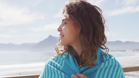 Portrait-of-happy-mixed-race-woman-standing-by-the-sea-with-blanket-over-shoulders-smiling