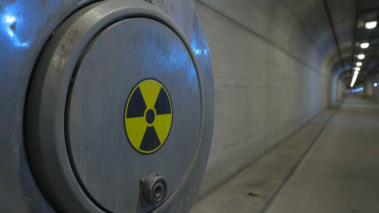 Close view of weathered metal door with black and yellow radiation trefoil remains nearly steady as concrete passage extends into depth beneath a regular line of ceiling lights.