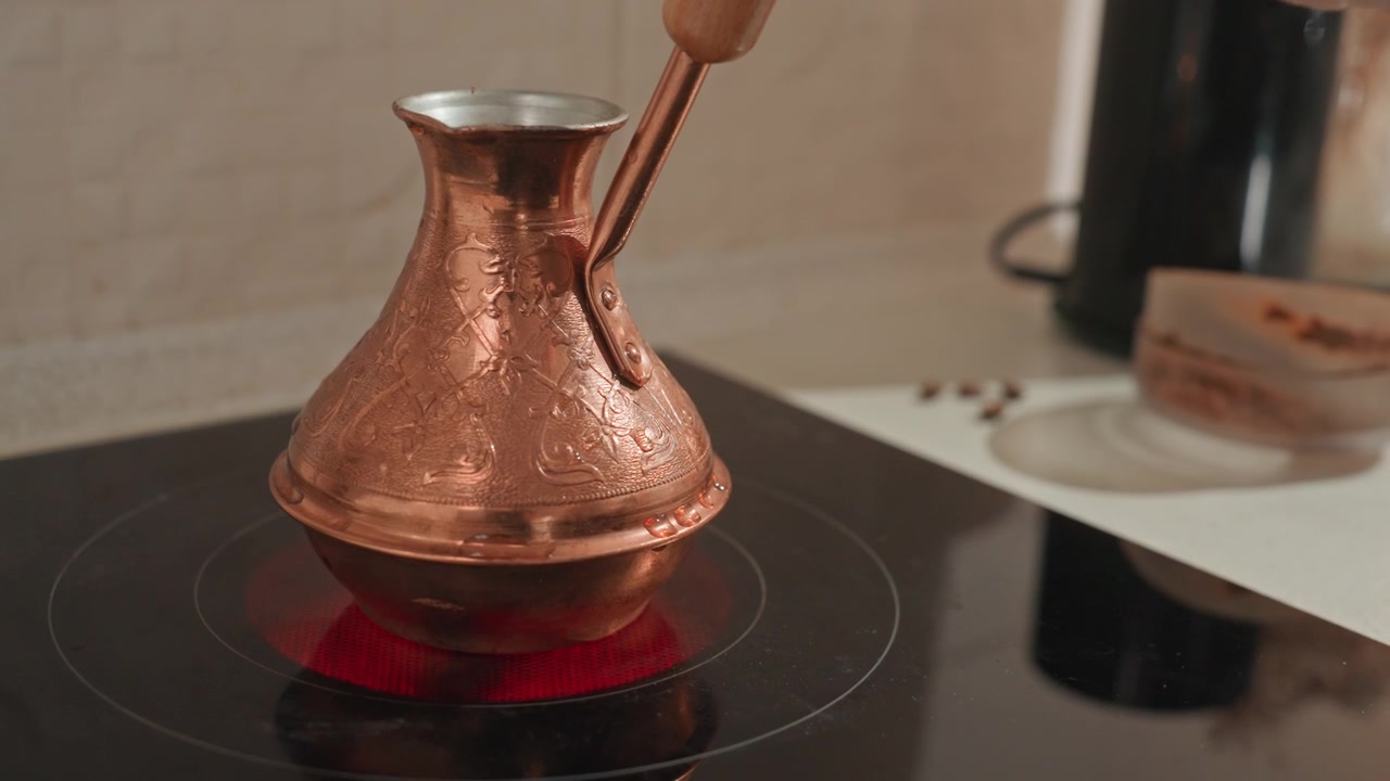 Nærbillede af kobber cezve på komfur, hånd rører mørkt kaffegrums og skum, rødt varmeelement gløder, hamret metaltekstur, traditionel kaffetilberedning, stille køkkenbordsscene, varm damp