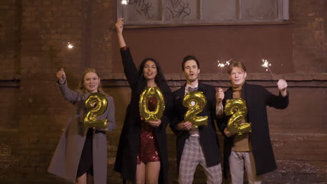 grupo de amigos vestidos con ropa elegante sosteniendo bengalas y 2022 números globos dorados en la calle después de la fiesta de año nuevo 1