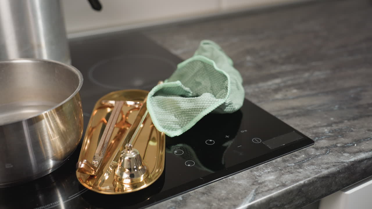 Gas cook top with golden tray and matching golden knife placed beside stainless pot, as handkerchief is dropped during food prep in modern kitchen setting with black glass surface and clean decor