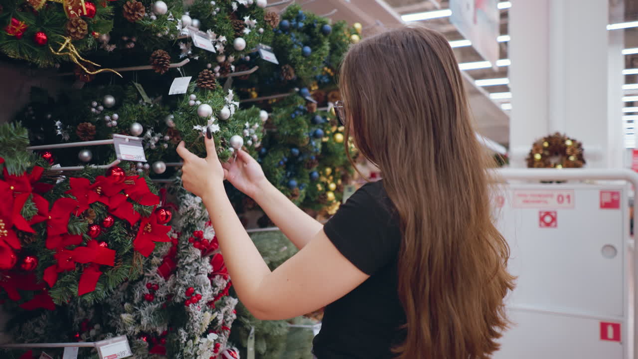 mujer joven con gafas y cabello marrón largo en camisa negra navegando por coronas festivas decoradas con adornos y piñas en la sección de vacaciones de la tienda con luces brillantes y señal direccional roja