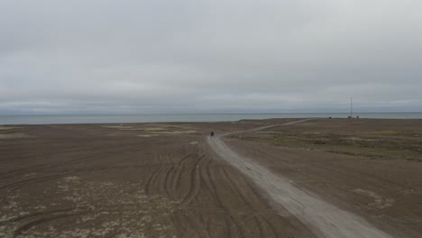 Toma-Aérea-De-Drones-De-Seguimiento-De-Un-Hombre-Solitario-En-Atv-En-La-Playa-De-Arena-Negra-Y-El-Océano-ártico-En-El-Punto-Más-Septentrional-Del-ártico-Estados-Unidos-Cerca-De-Barrow-Alaska