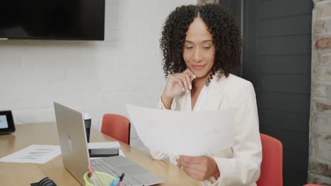 una mujer de negocios biracial sonriente en el escritorio con una computadora portátil sosteniendo un documento en la oficina, en cámara lenta.