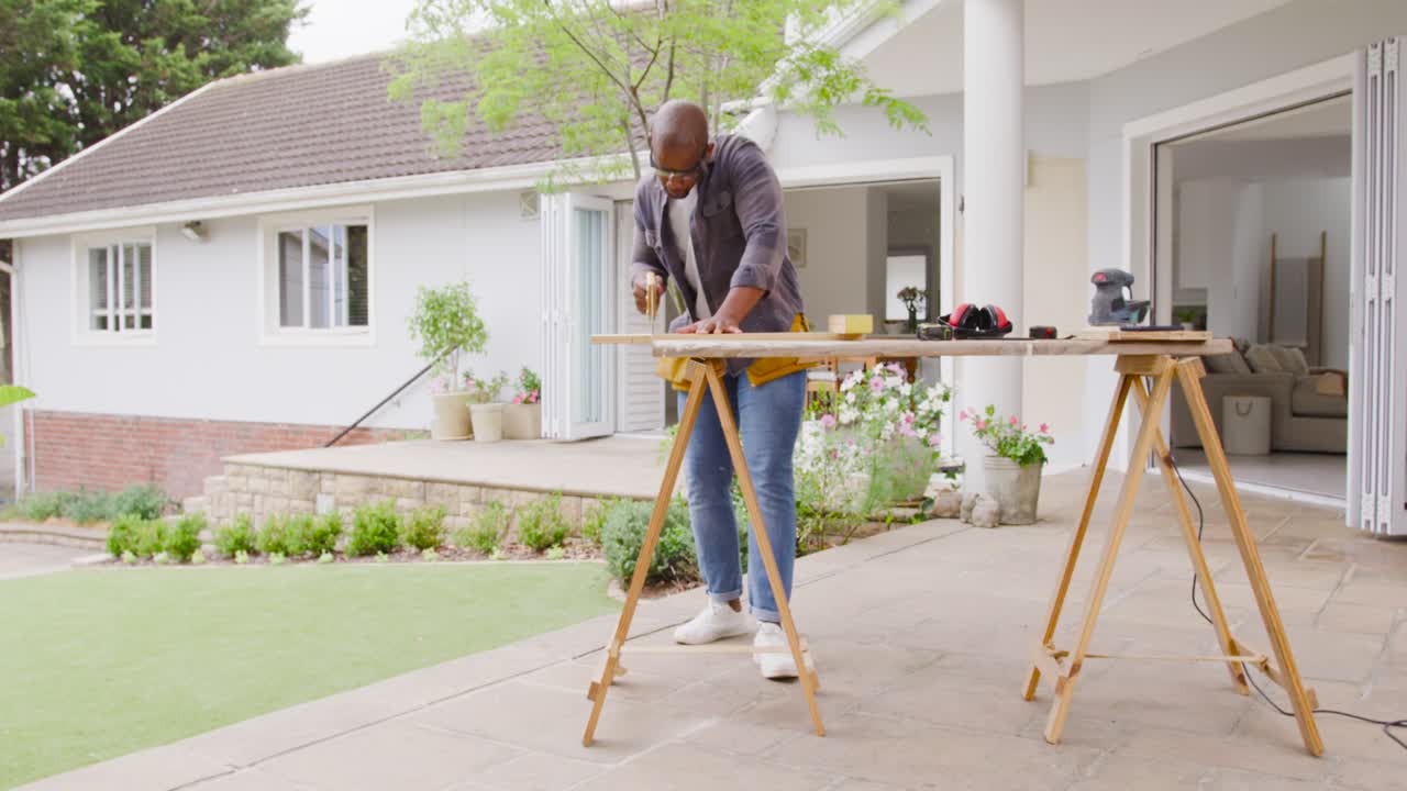 Après avoir placé une planche sur des tréteaux sur la terrasse, un homme afro-américain prend des mesures pour préparer la prochaine coupe.