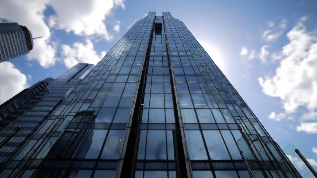 Witness the beauty of modern skyscraper design as sunlight reflects off glass facades. The towering building stands majestically against a backdrop of scattered clouds, showcasing urban innovation.
