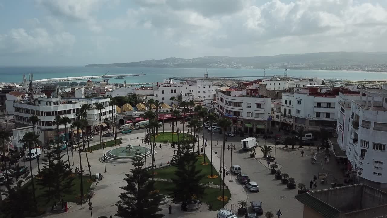 Vista aérea cinematográfica de uma praça pública movimentada em Tânger, Marrocos, com edifícios brancos, palmeiras e o litoral mediterrâneo ao fundo