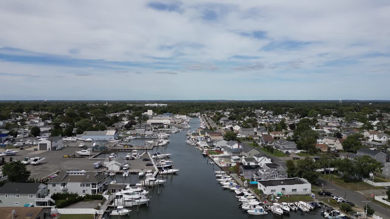 Droneutsikt som flyr over en vannvei omgitt av bolighus og en båthavn med mange fortøyde yachter, som representerer en velstående forstadslivsstil en delvis skyet sommerdag