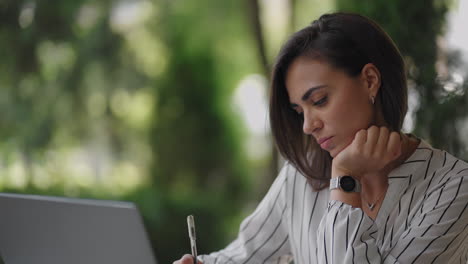 mujer de negocios morena de etnia hispana trabaja remotamente mientras está sentada en un café de verano en un día soleado con una computadora portátil y escribe con un bolígrafo y un cuaderno