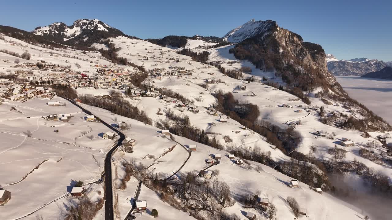 Aerial view of a winding road leading through a picturesque winter landscape in Amden, Switzerland, with snow-capped mountains and a sea of fog in the distance.
