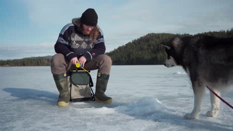 man ice fishing with his alaskan malamute dog in the frozen lake