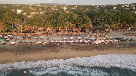 Drone-Acercándose-A-La-Punta-En-La-Playa-De-Zicatela-Puerto-Escondido-Costa-De-Oaxaca-En-México-Con-Gente-Tomando-El-Sol-En-Una-Playa-Tropical-De-Arena-Con-Palmeras-Y-Paraíso-Turístico