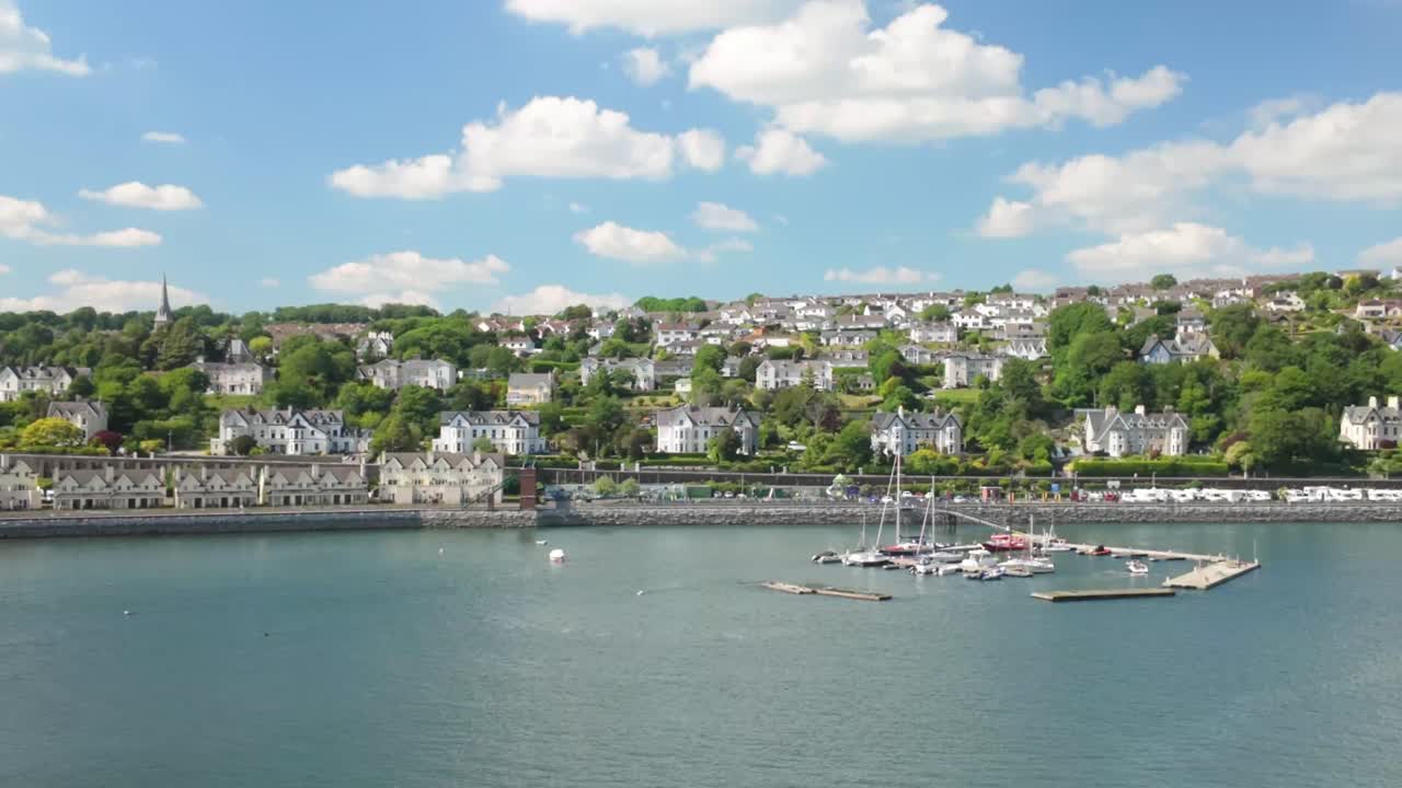 Slow-moving cruise ship passes a quiet coastal section of Cork, Ireland. The scene features small seaside houses, a modest pier, and calm water under sunny skies