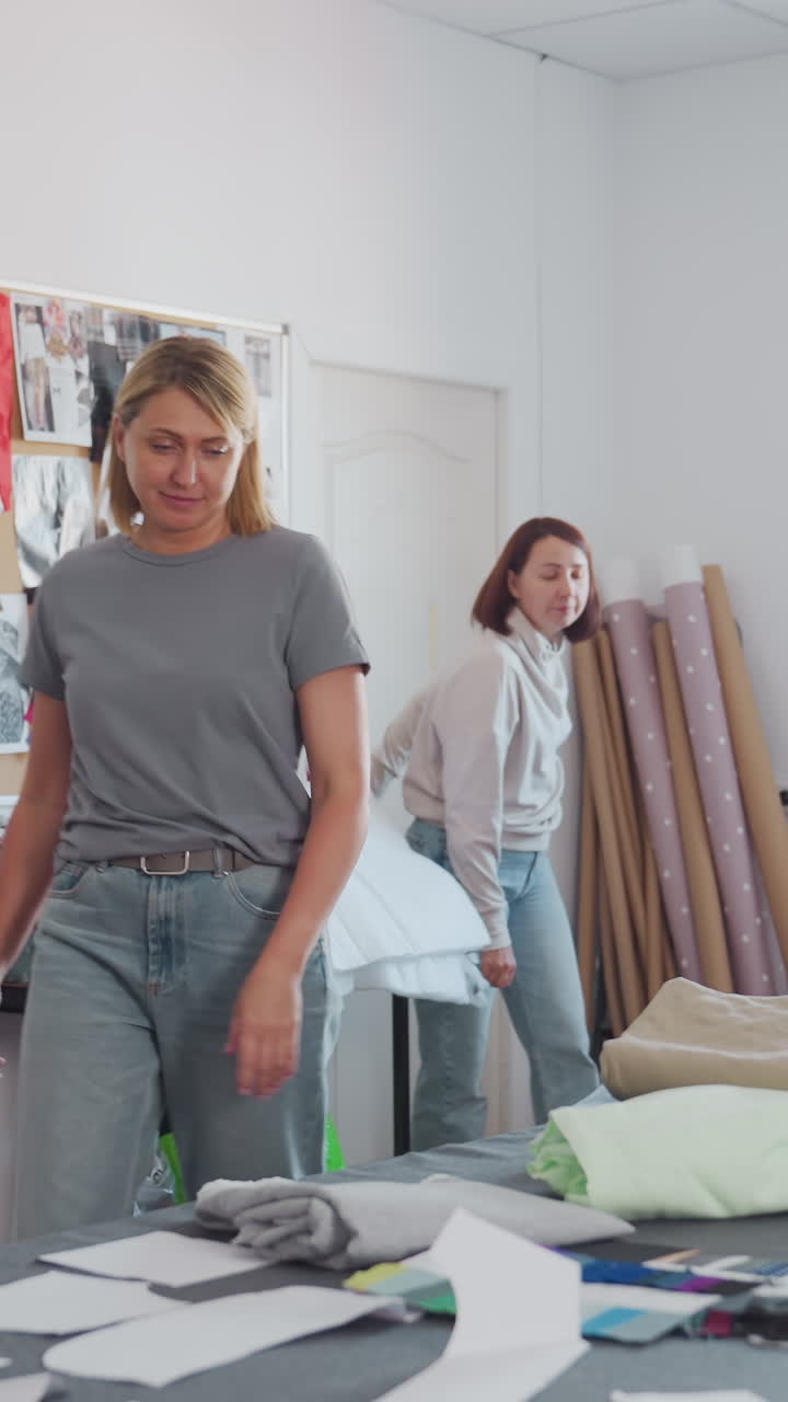 Planificación de estudio con telas y patrones. Dos diseñadoras revisan piezas de patrones sobre la mesa de corte, doblan muestras, señalan bocetos, luz brillante de la ventana, energía colaborativa, consulta con el cliente.