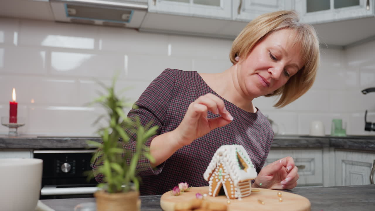 frau, die das gingerbread-haus sorgfältig mit grünen perlen dekoriert, eine gefallene perle aufnimmt und zurücklegt, gemütliche küchenumgebung mit kerze, topfpflanze, holzbrett und warmer beleuchtung