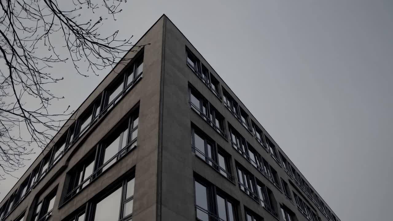 Low angle view of a gray concrete building with numerous dark windows, emphasizing stark urban geometry against a cloudy sky, with bare tree branches adding a touch of nature to the urban scene