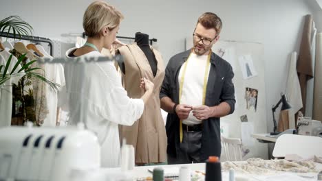 Dos-Jóvenes-Diseñadores-De-Ropa-Con-Estilo,-Hombre-Y-Mujer,-Trabajando-Juntos-Sobre-El-Vestido-Con-Cintas-Métricas-Y-Hablando-En-El-Estudio