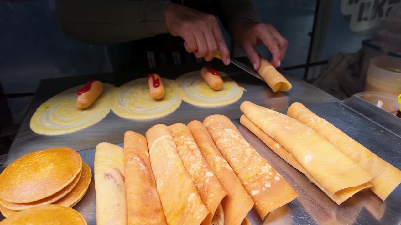 Primo piano di un venditore che arrotola frittelle sottili con salsiccia su una griglia metallica per street food.