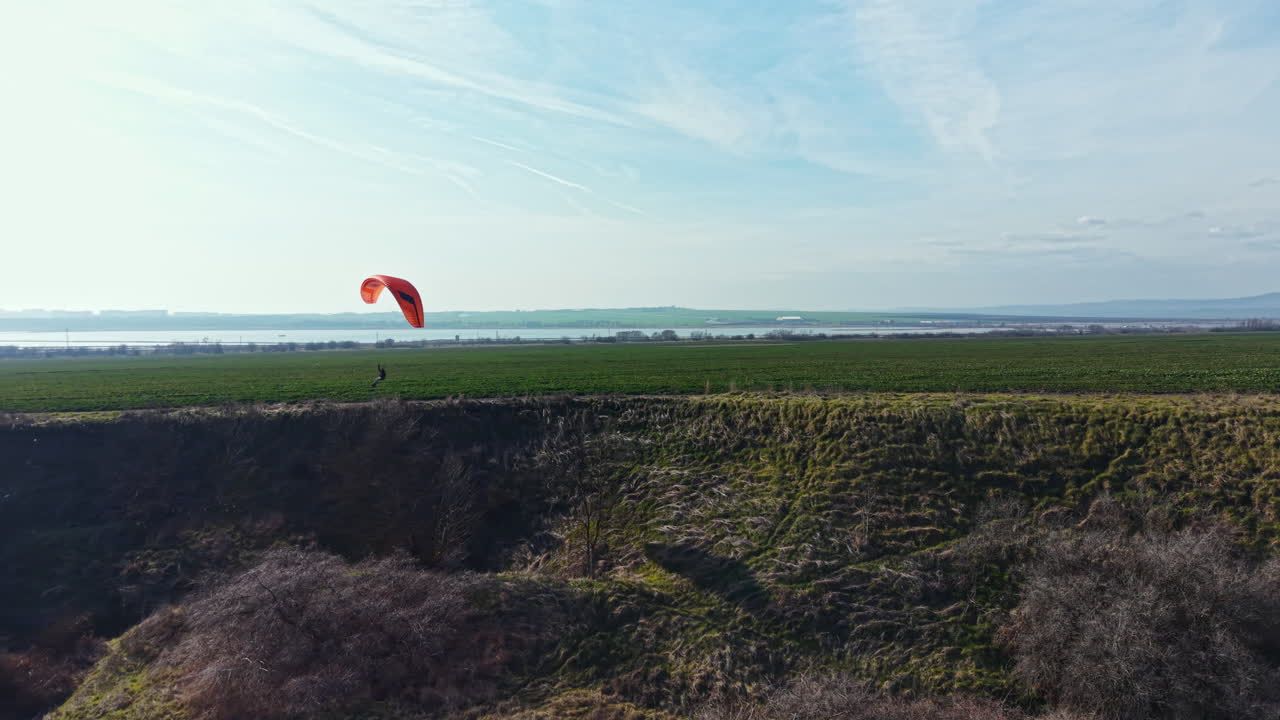 Pessoas praticam parapente sobre campos verdes perto de um grande corpo d'água num dia claro, numa área rural com céus abertos.