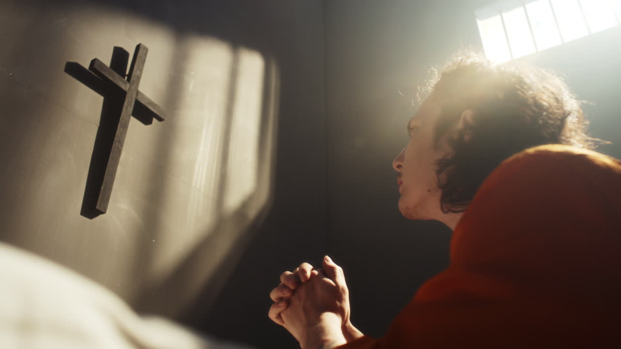 Inmate Clasping Hands and Praying in Prison Cell with Cross Mounted on Wall