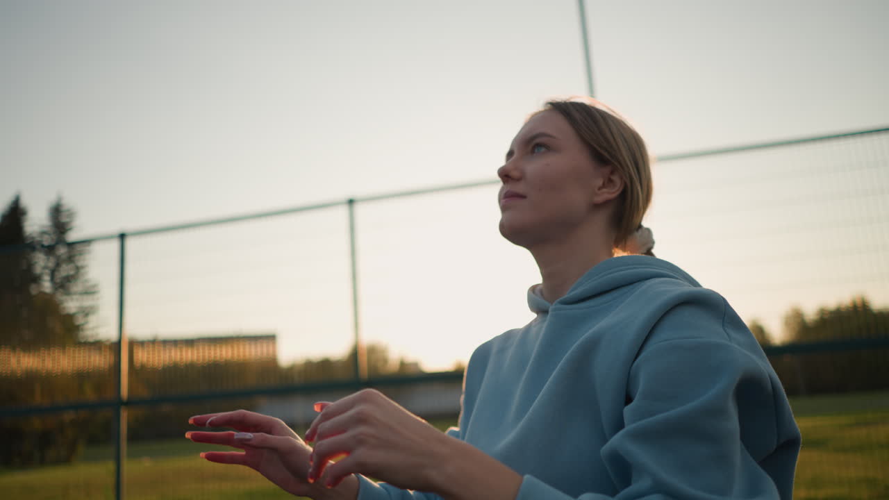dama con suéter cian jugando al voleibol con el enfoque en sus manos, al aire libre con cielo brillante y valla en el fondo, activa y comprometida en la práctica de voleibol bajo la luz solar natural