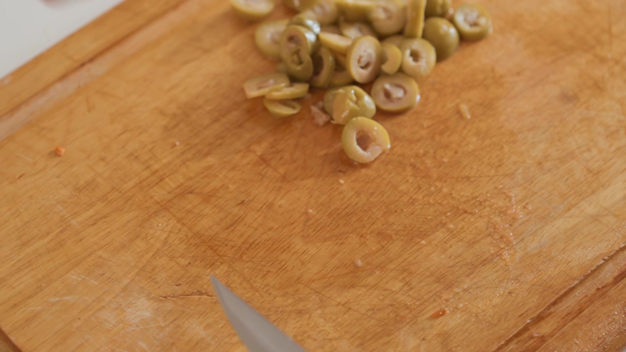 Scena di cucina in primo piano che mostra una mano che affetta piccole olive verdi a rondelle sottili, poi raccoglie i pezzi in un mucchietto ordinato su un caldo tagliere di legno con una luce soffusa dall'alto, con mano, coltello, taglio.