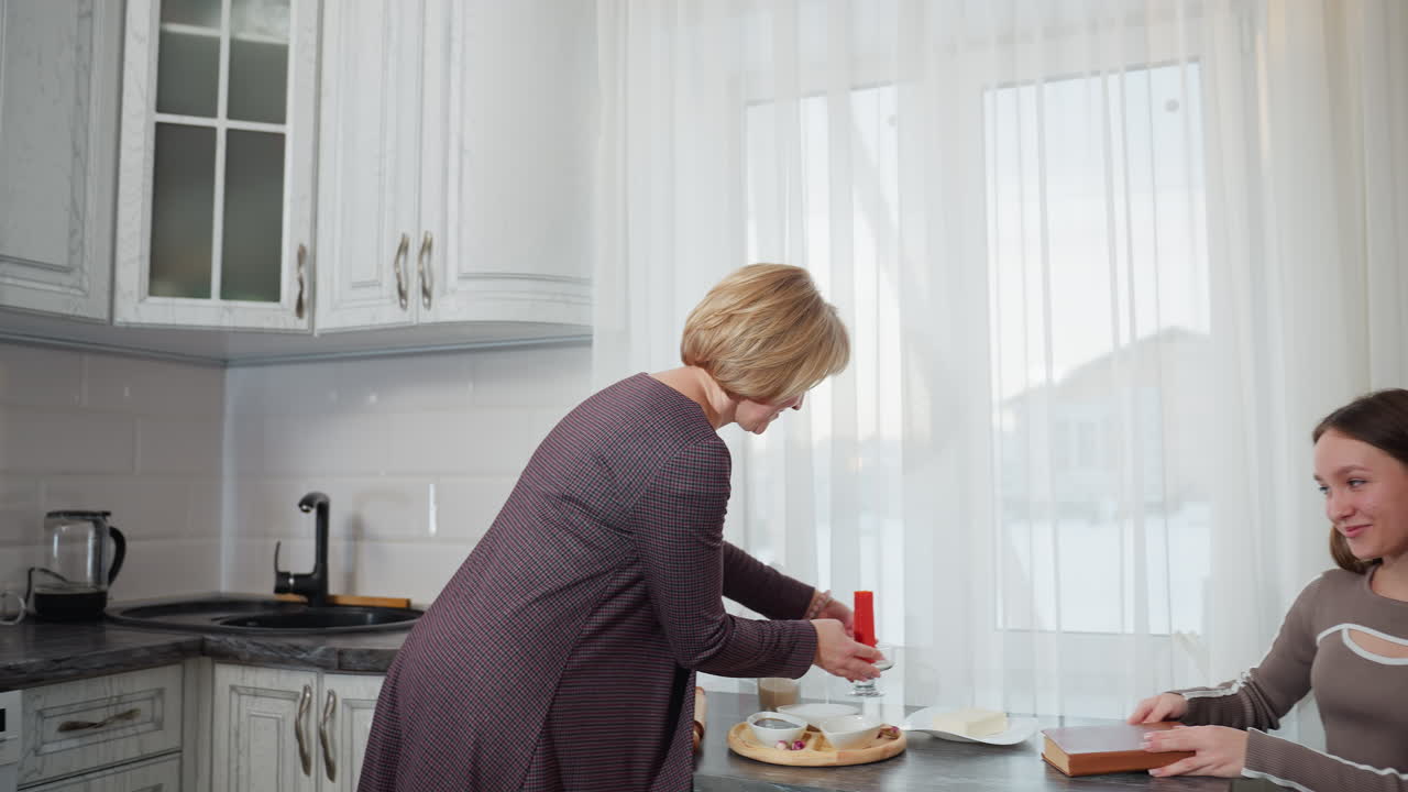 여성은 현대 부에서 은 불을 들고, 딸이 앉아서 읽는 테이블에 놓을 준비를 하고 있습니다. 부 장비와 장식은 밝은 자연 조명 속에서 쾌적한 가정 분위기를 만니다.