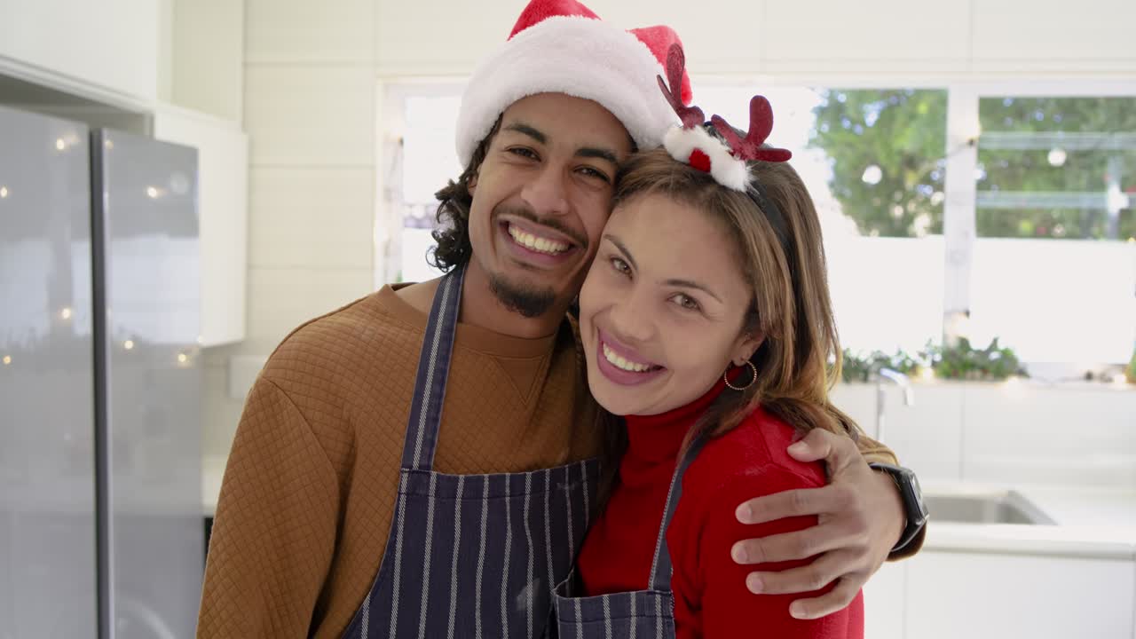 Pareja diversa con delantales y gorros navideños bailando en la isla de la cocina después de hornear galletas