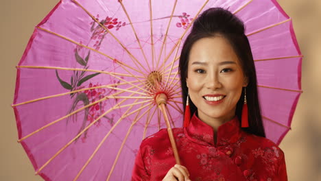 close-up view of asian young cheerful woman in red traditional clothes holding parasol while smiling at camera