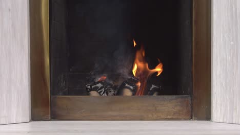 the logs burn down in fireplace, emitting a light smoke and creating warmth and comfort in the house, front view. piece of furniture in home interior, living or dining room