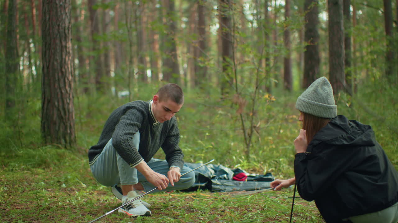 Couple outdoors camping in forest as man collects tent pole from woman who hands it over while standing near scattered gear and tent fabric on grassy woodland floor under soft daylight