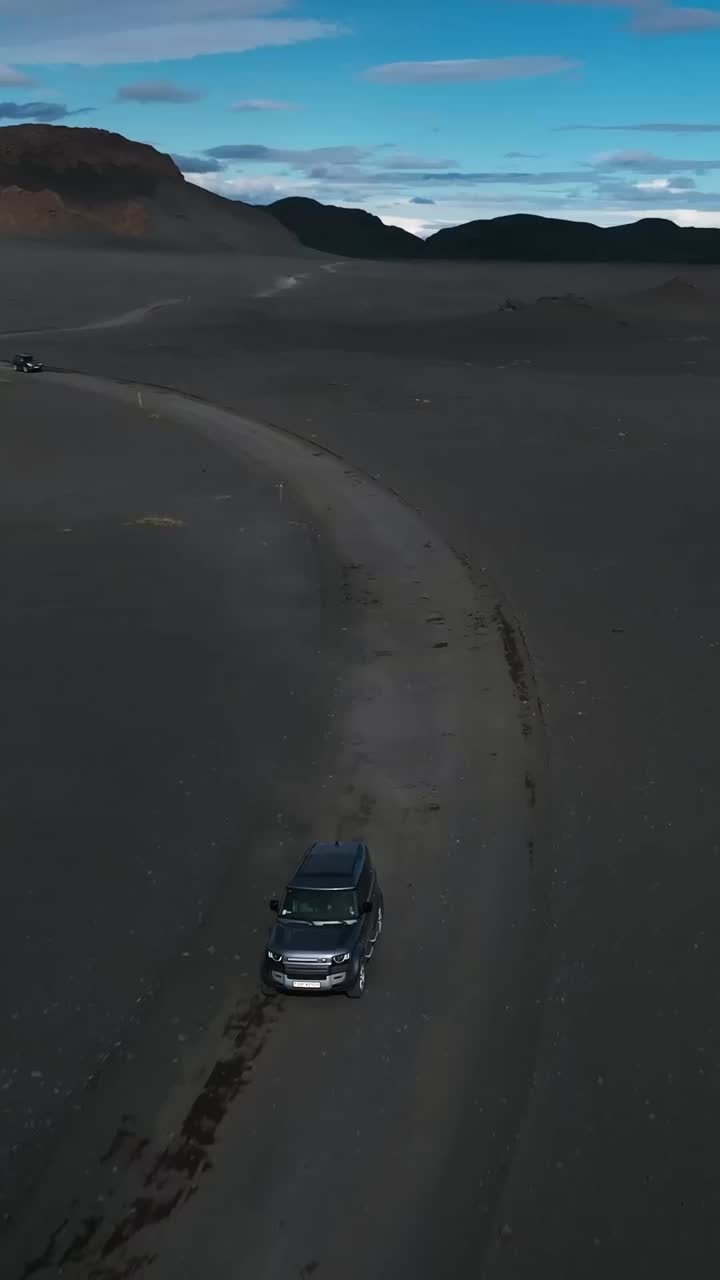Aerial view of an SUV driving along a winding gravel road through Iceland’s dark volcanic landscape, with rugged hills and scattered clouds in the background