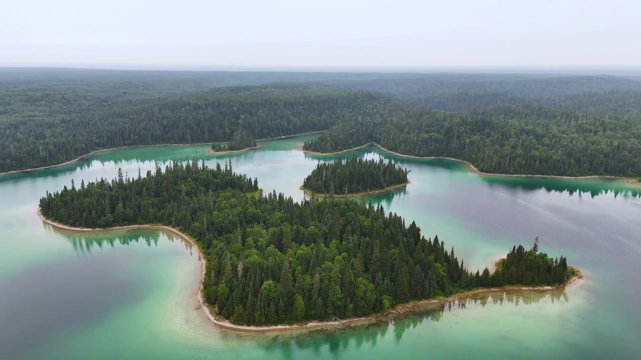 Luftfoto af en skovklædt ø med en buet sandbugt omgivet af turkis og grønt vand ved Arnott Lake i det nordlige Ontario